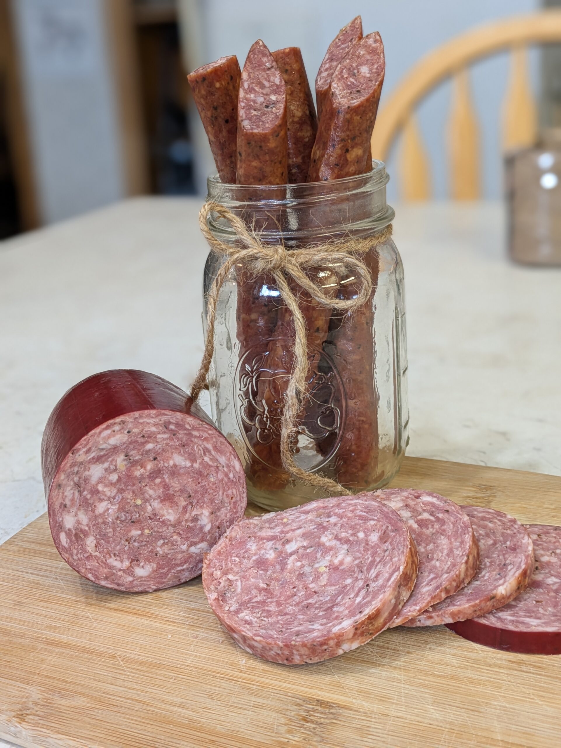 Homemade cured sausage sticks in a mason jar with a twine bow, alongside sliced rounds of sausage on a wooden cutting board, showcasing a rustic food presentation.