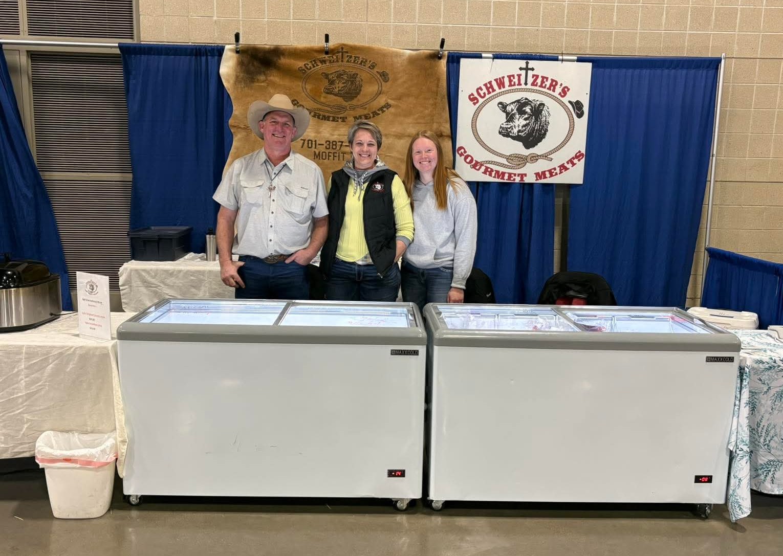 Tara Splonskowski - Vendor Show A family team representing Schweitzer's Gourmet Meats stands at their booth featuring two display freezers filled with various meat products, showcasing their offerings at a local event.