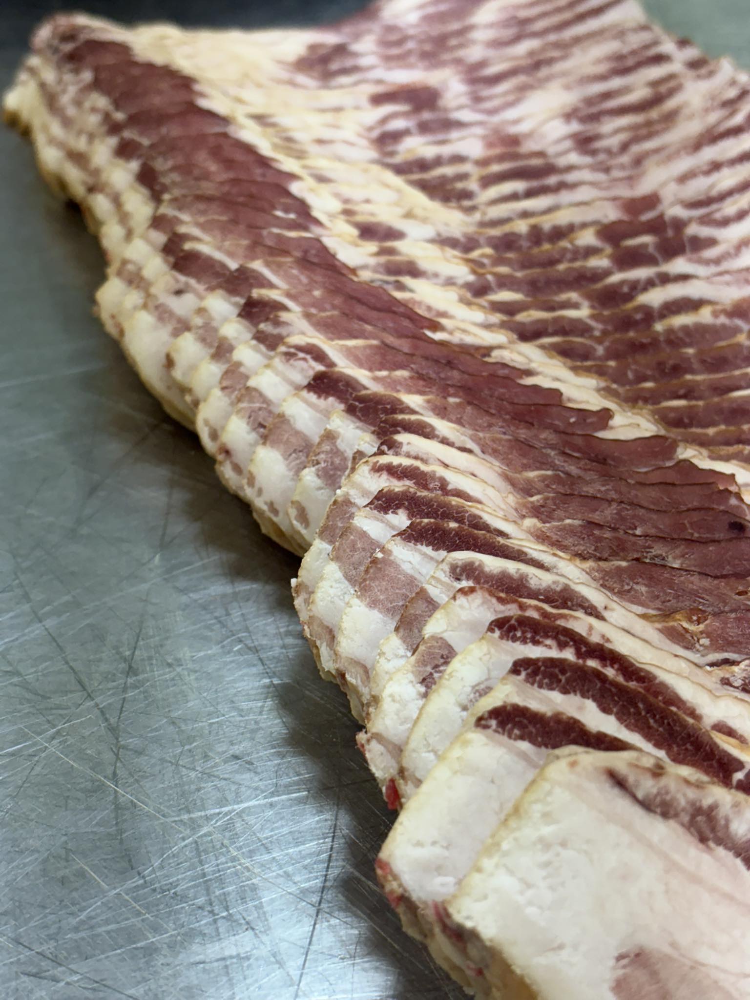 Close-up view of freshly sliced bacon strips showcasing layers of meat and fat on a stainless steel surface, ideal for culinary presentations and recipes.