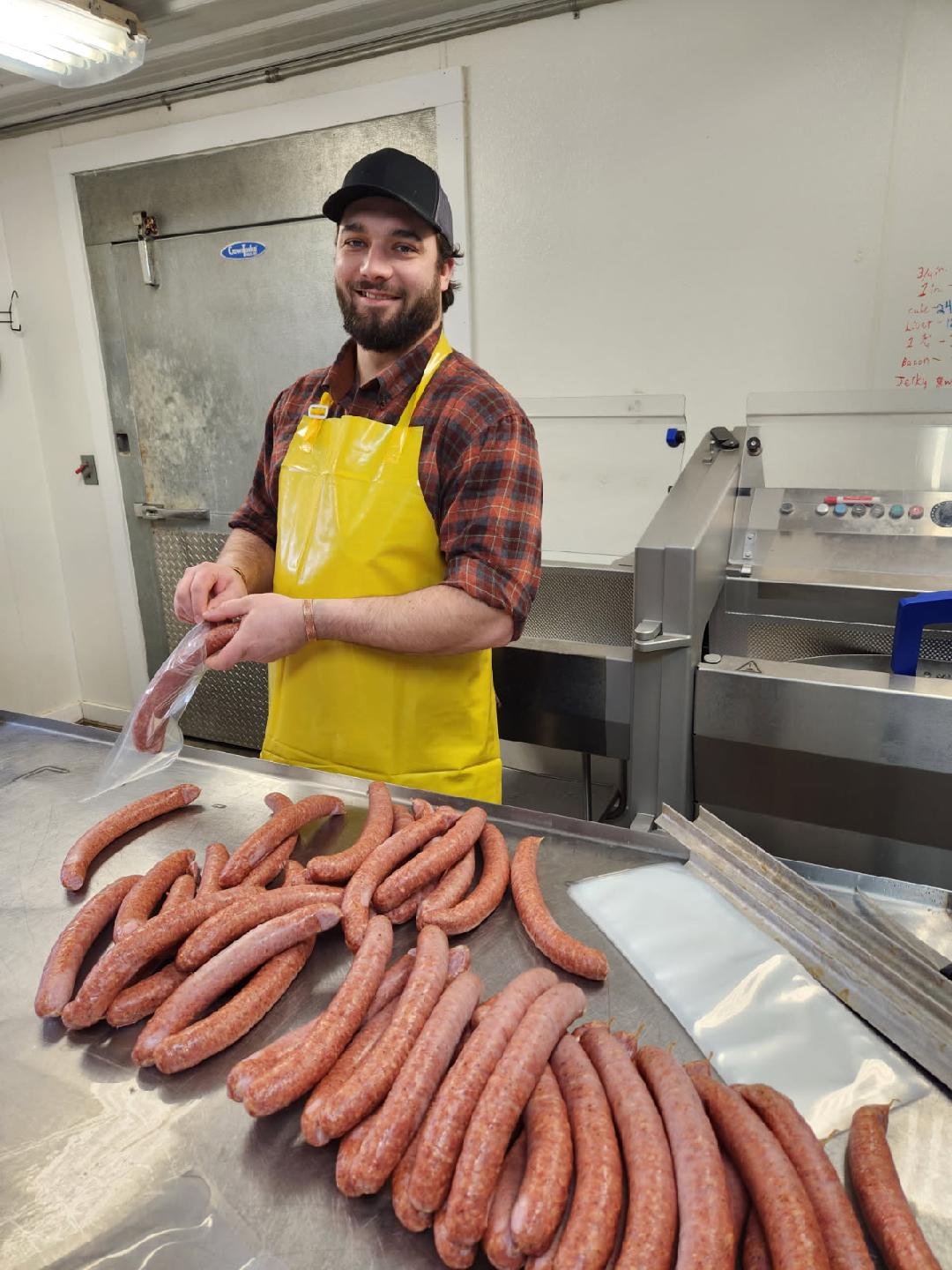 Smiling butcher in a yellow apron preparing sausages in a clean meat processing facility, with freshly made sausages arranged on a stainless steel table.