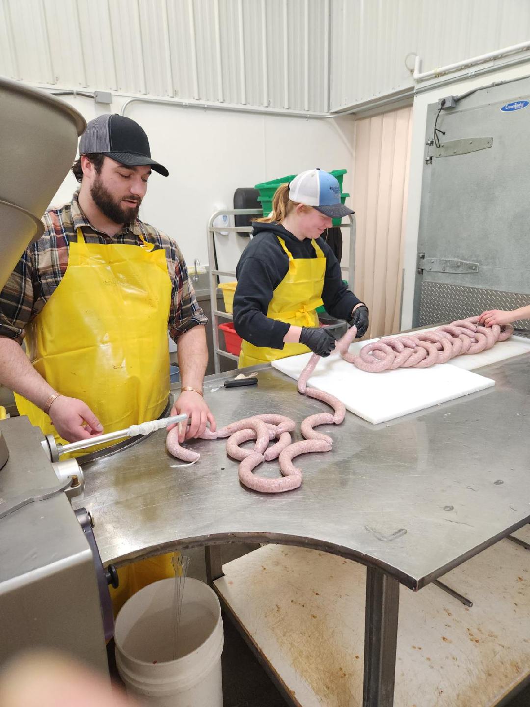 Tara Splonskowski - image000002_20260205_095009 Two individuals in yellow aprons are preparing sausage in a processing facility. One person is operating a sausage stuffer while the other shapes and ties the sausage links on a stainless steel table. Colorful bins are visible in the background, indicating a busy meat processing environment.
