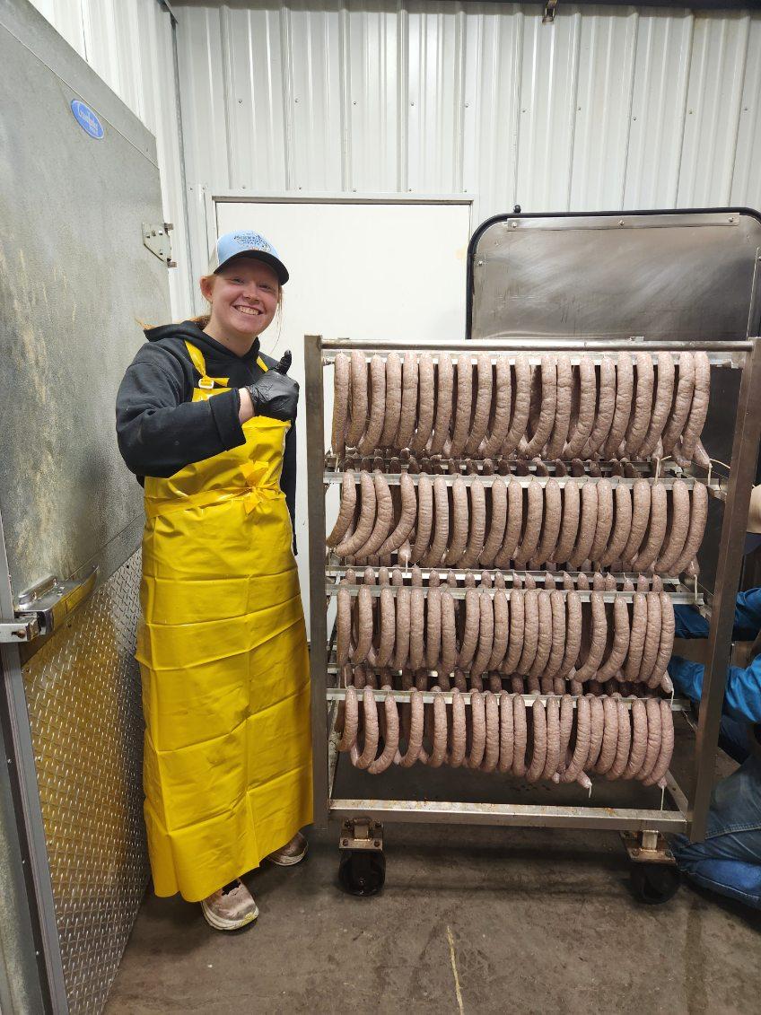 Tara Splonskowski - image000005_20260205_095009 Smiling worker in a yellow apron and gloves stands beside a rack of freshly made sausages in a processing facility, showcasing the sausage production process.