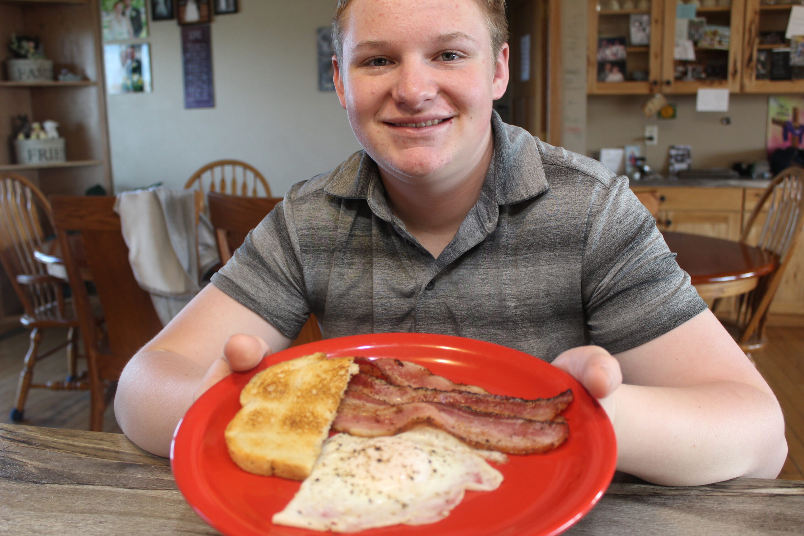 Tara Splonskowski - IMG_6435 A smiling teenager holds a red plate featuring a hearty breakfast of crispy bacon, a fried egg, and toasted bread, set against a cozy kitchen background.