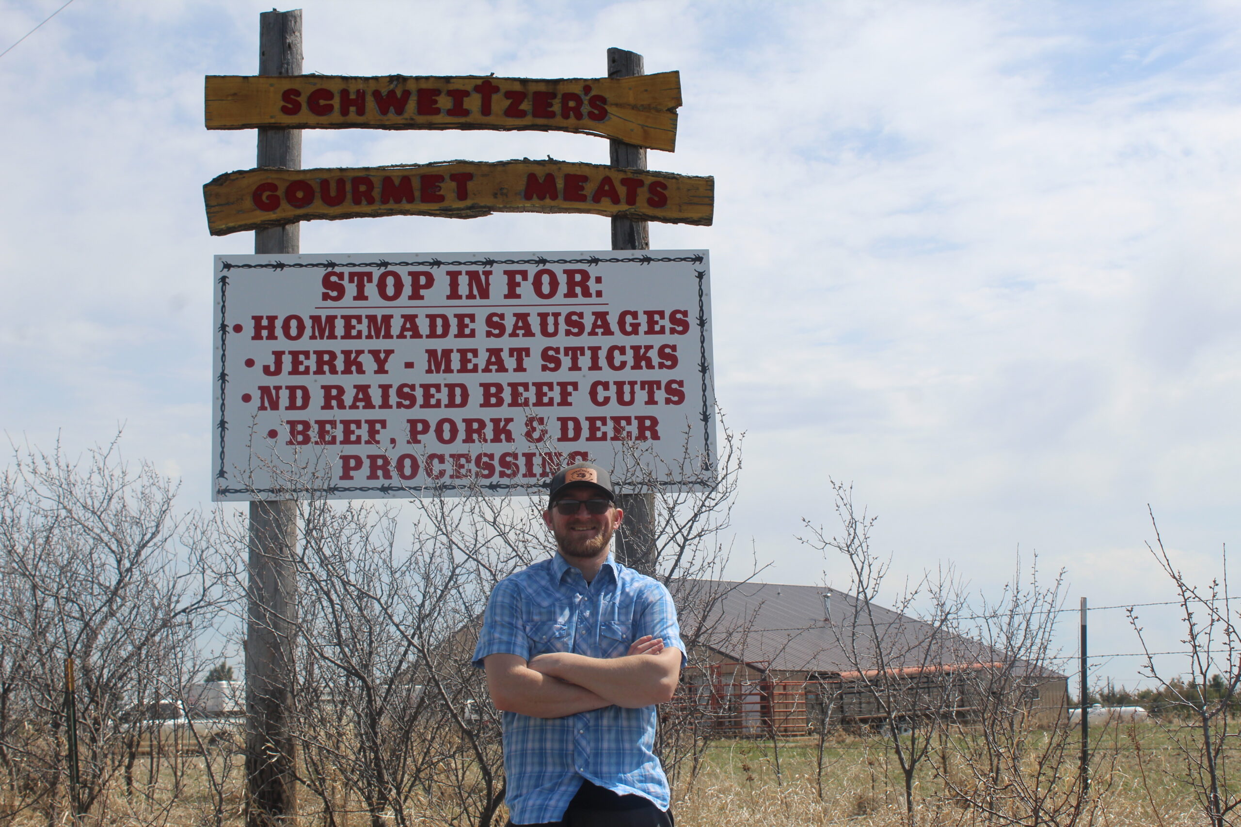 A man stands in front of the Schweitzer's Gourmet Meats sign, advertising homemade sausages, jerky, meat sticks, North Dakota raised beef cuts, and beef, pork, and deer processing services. The background features a cloudy sky and a rustic building.