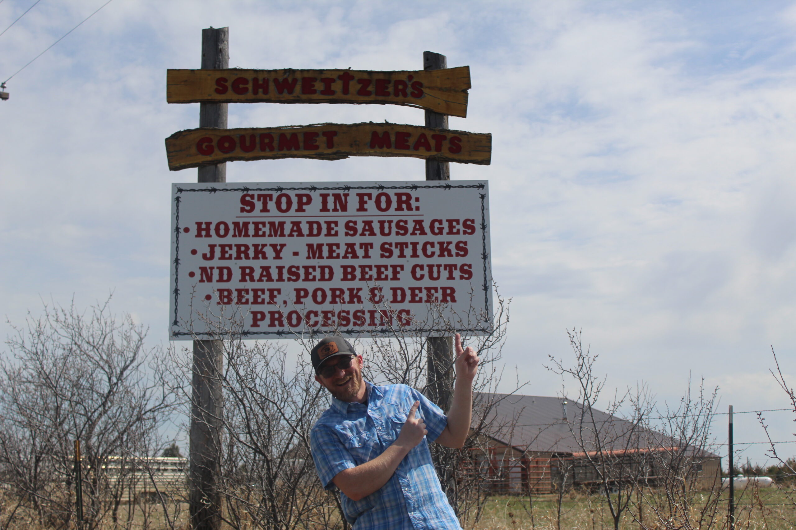 Tara Splonskowski - IMG_6439 Sign for Schweitzer's Gourmet Meats advertising homemade sausages, jerky, meat sticks, North Dakota raised beef cuts, and beef, pork, and deer processing services, with a smiling man in a blue shirt pointing at the sign.