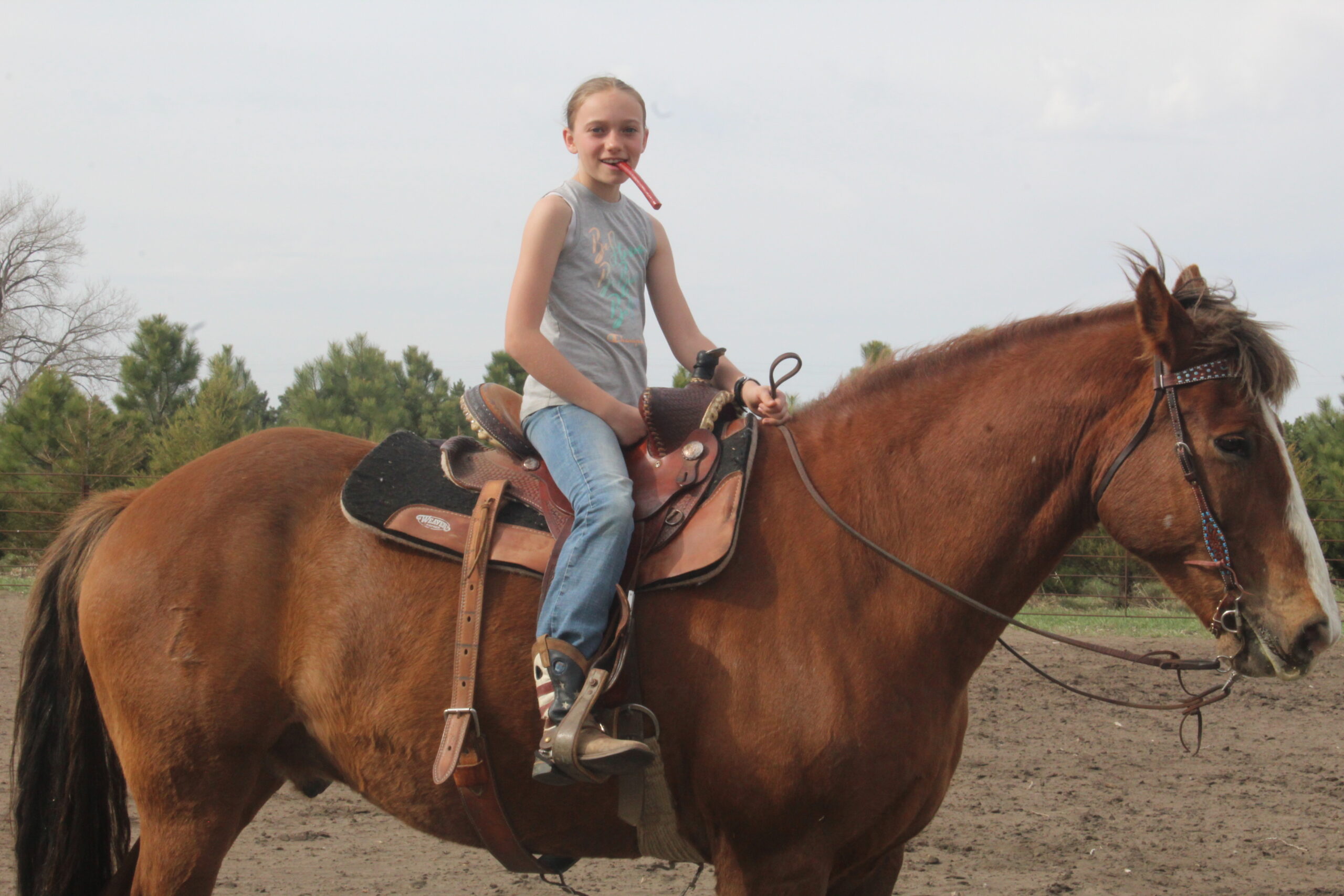 Tara Splonskowski - IMG_6441 Young girl riding a brown horse in a saddle, smiling and holding a stick in her mouth, with a rural landscape and trees in the background.